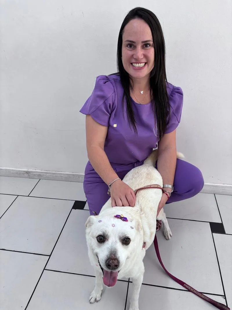 M.V Roberta agachada ao lado de uma cadela idosa branca, sorrindo, com a mão apoiada nas costas dela